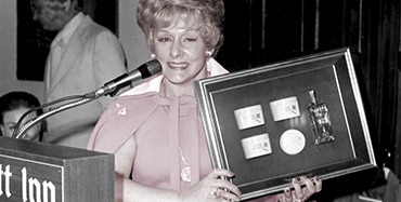 Mary Kay Ash smiles for a photo while standing at a podium and holding up a plaque.