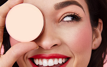 Brunette woman smiling and holding Mary Kay product playfully over her eye.d.
