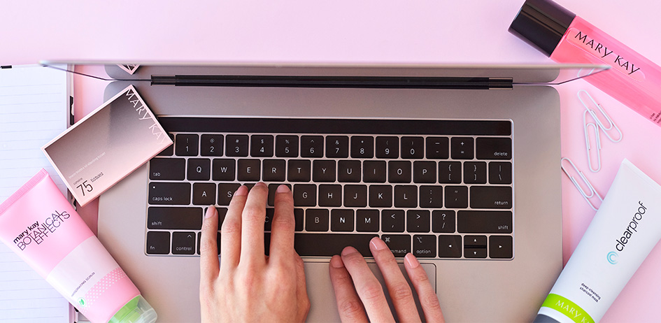 Woman’s hands typing on a laptop computer surrounded by Mary Kay products and bright, colorful graphics.